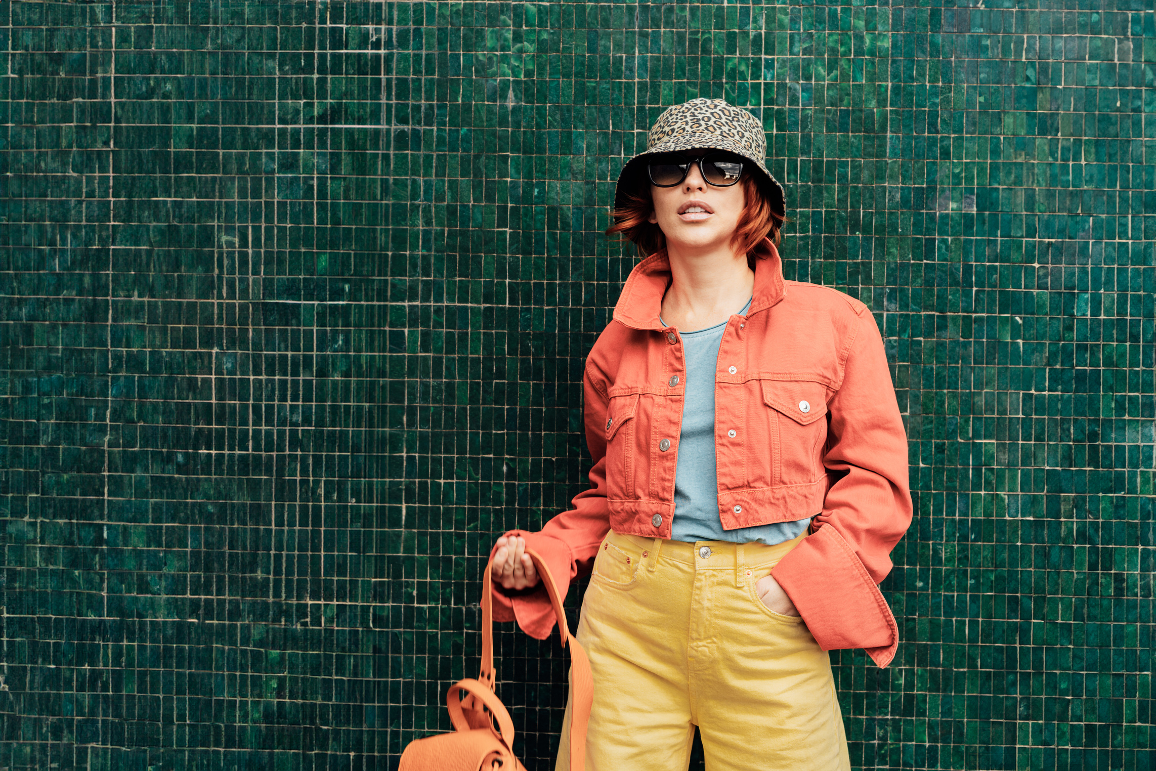 Hipster young woman in trendy peach color jacket, sun glasses, backpack bag and bucket hat posing on the green tile wall background. Color of the 2024 year. Urban city street fashion. Copy space